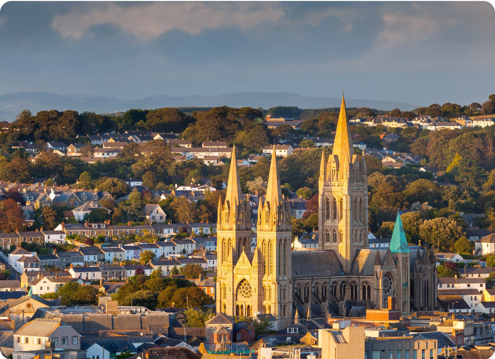 A scenic view of Truro, Cornwall, featuring the impressive Truro Cathedral with its three striking spires standing tall amidst a backdrop of houses, trees, and distant hills under a cloudy sky. The evening light casts a warm glow on the cathedral and surrounding area.