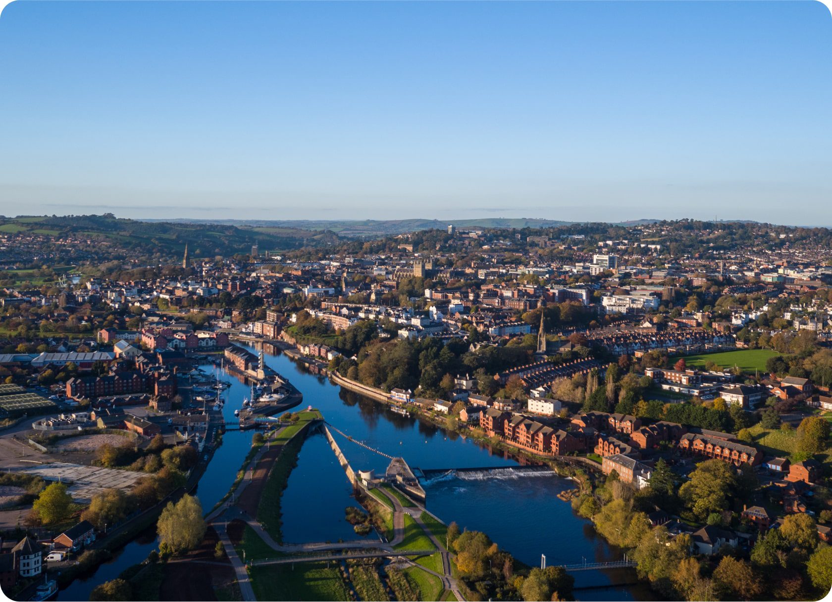 Aerial view of a city with a river winding through it. The river is surrounded by greenery and buildings, including residential and commercial areas. The landscape is illuminated by daylight, under a clear blue sky. Several bridges span the river, connecting both sides.