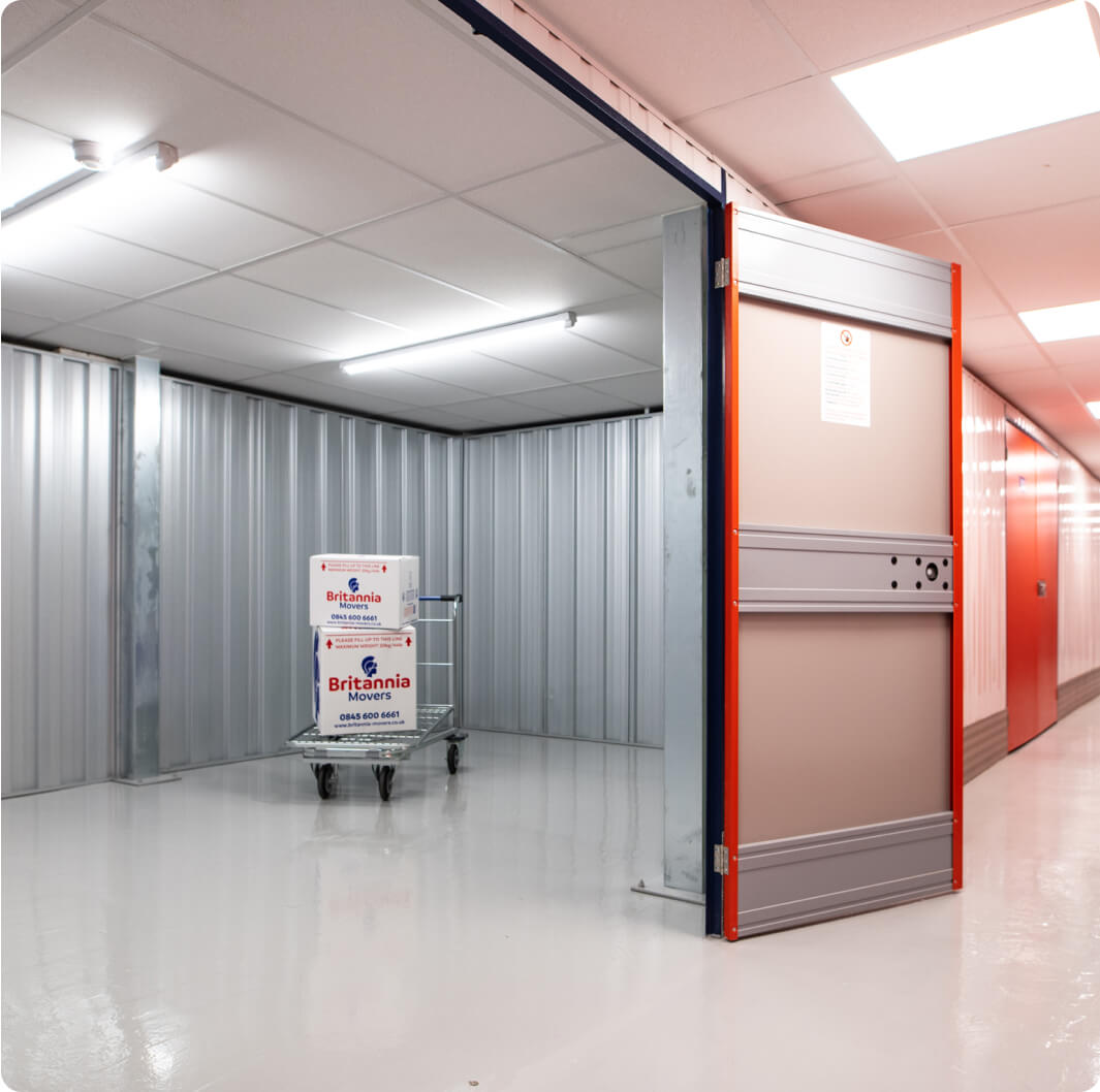 A storage unit with an open door revealing a clean, well-lit interior. Inside, a metal cart holds two stacked cardboard boxes labeled "Britannia" on a shiny white floor. The walls and ceiling are made of metallic panels, and the aisle has bright lighting.