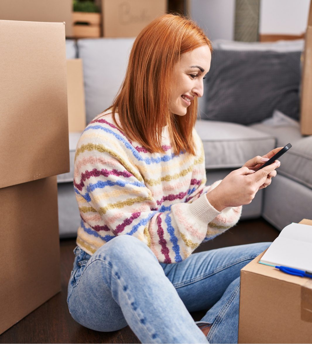 A person with red hair sits on the floor among moving boxes, smiling while looking at a smartphone. They are wearing a striped sweater and jeans. An open notebook rests on a box beside them. The background features a couch with cushions.