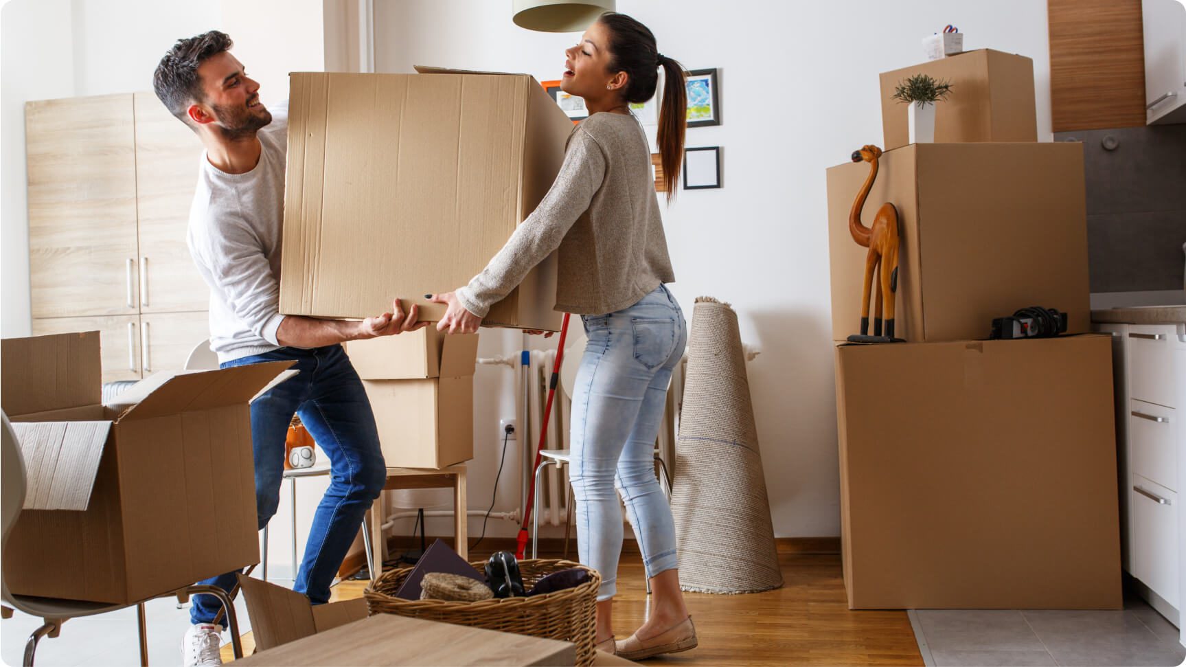 A man and woman are in a modern apartment, lifting a large cardboard box together. The space is filled with moving boxes and household items, including a wrapped rug and a decorative giraffe statue. The woman is smiling, and the scene conveys a sense of teamwork and moving.