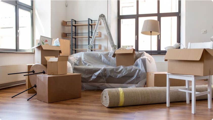 A room filled with moving boxes and household items. A couch covered in plastic sits in the center, surrounded by a ladder, stacked shelves, a rolled-up rug, a lamp, and scattered chairs. Large windows in the background let in natural light.