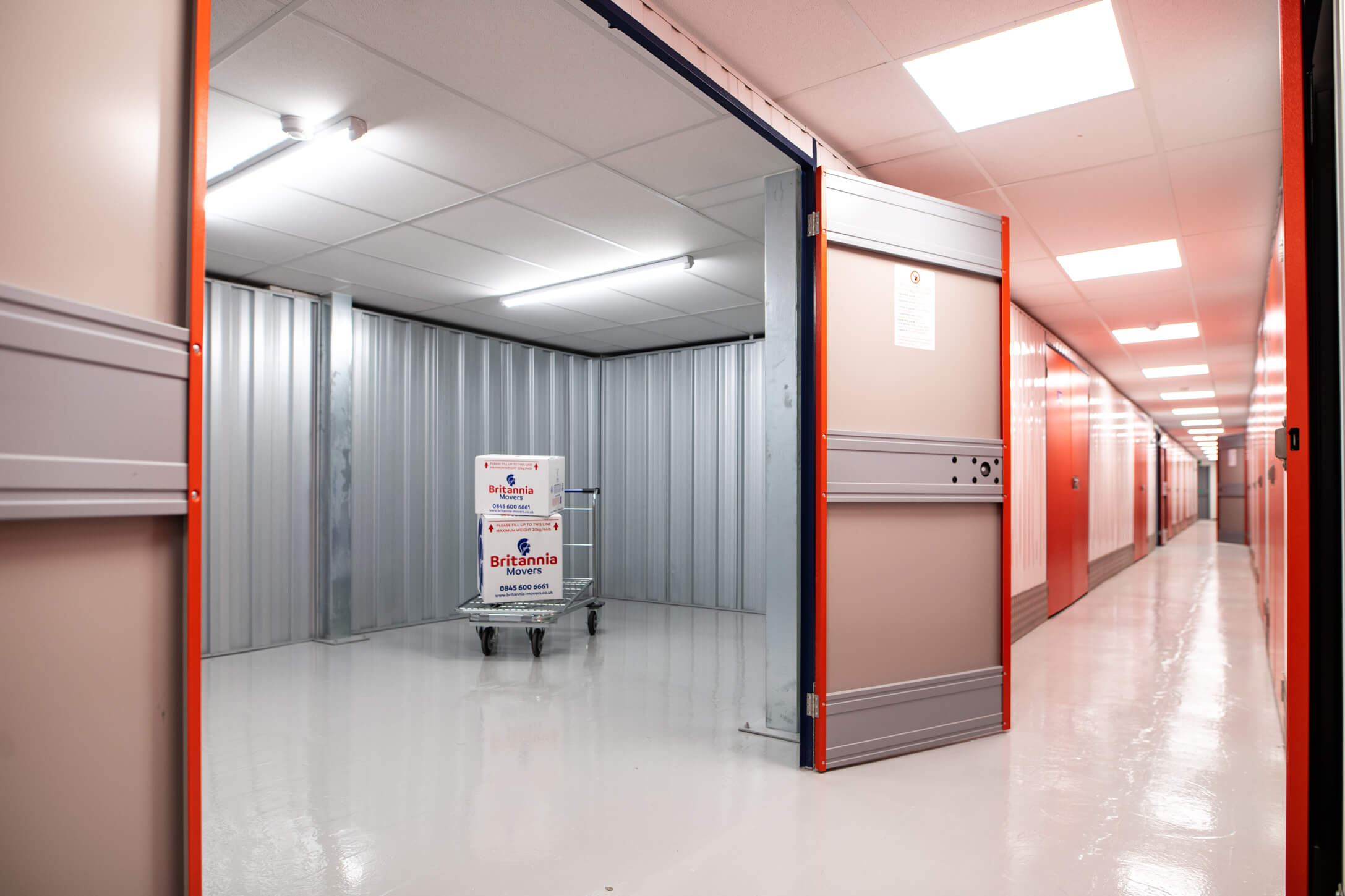 A brightly lit storage facility with a large open unit. The unit door is partially open, revealing a cart holding two stacked boxes inside. The facility has clean, polished floors and orange accents along the walls and doors.