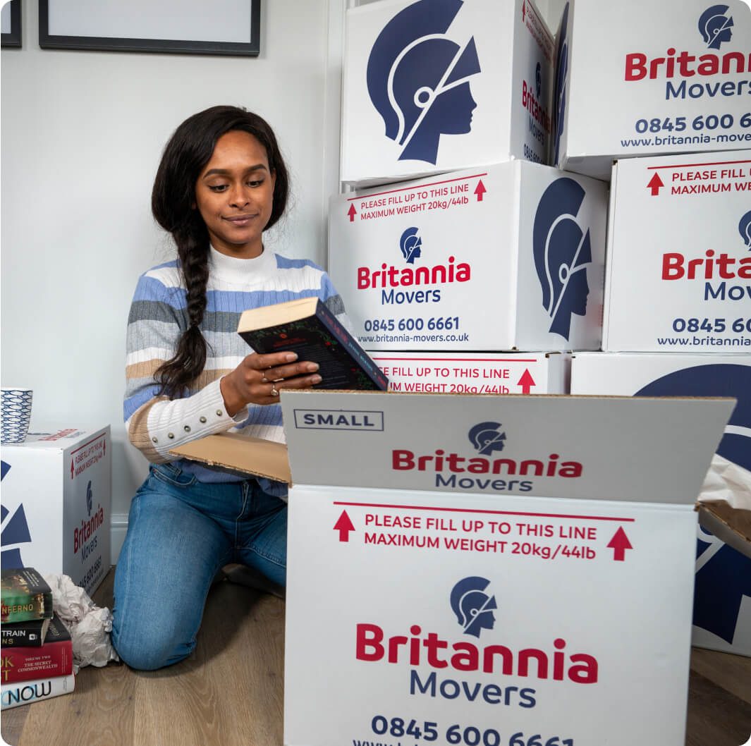 A woman kneels on the floor amidst several Britannia Movers boxes, packing a book into one of the boxes. She is wearing a striped sweater and jeans. The boxes are labeled with contact information and weight instructions.