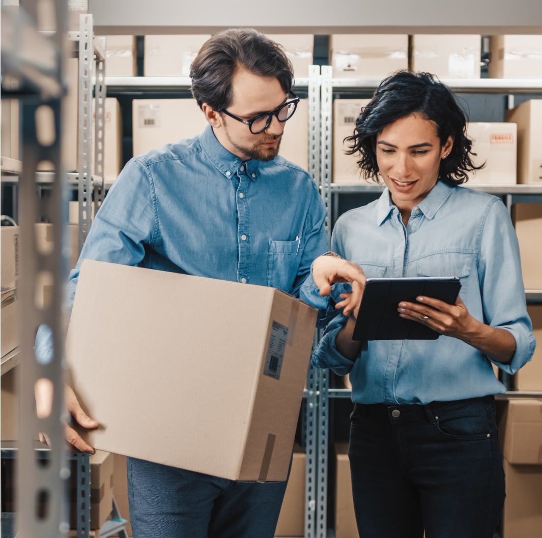 Two people in a warehouse, both wearing denim shirts. One person holds a cardboard box while the other points at a tablet screen. They are surrounded by metal shelves filled with boxes. They appear to be discussing or checking inventory.