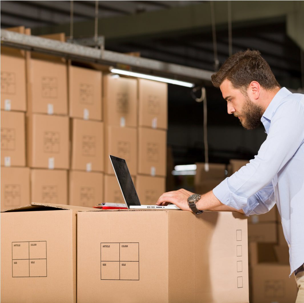 A man with a beard is working on a laptop placed on a stack of cardboard boxes in a warehouse. The background is filled with more stacked cardboard boxes, indicating a busy, organized storage environment. The man is focused, wearing a light shirt.