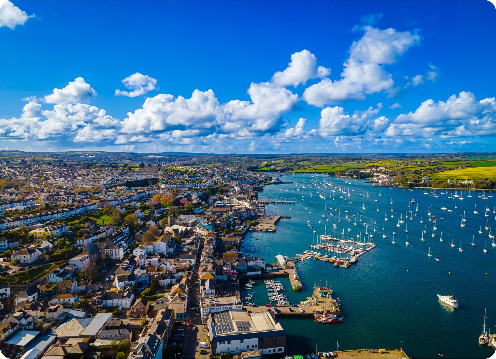 Aerial view of a coastal town with a harbor filled with boats. The town has numerous buildings, roads, and greenery. The sea is calm under a bright blue sky with scattered white clouds. Green fields are visible in the background.