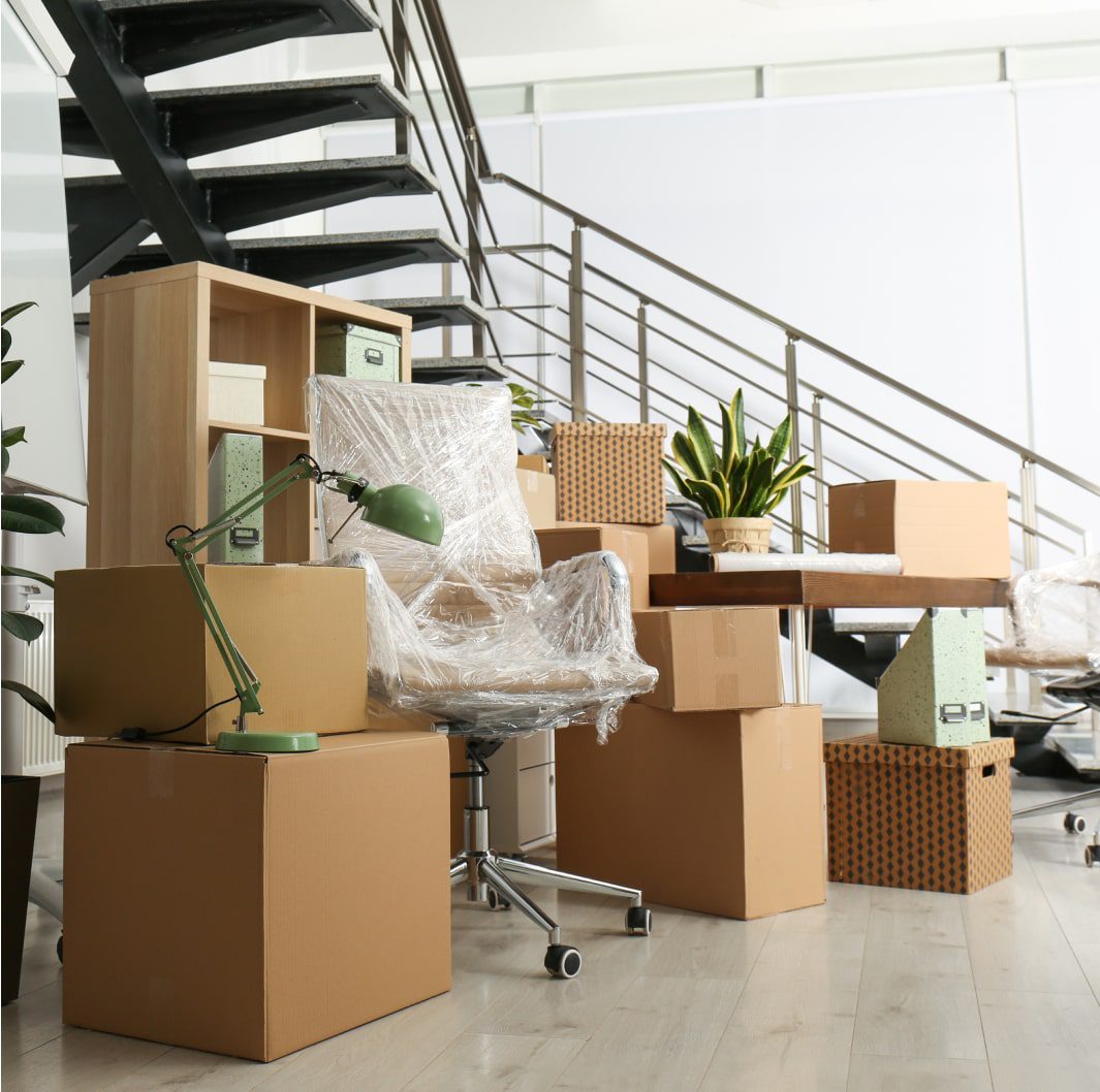 A modern office space filled with packed cardboard boxes, a wrapped office chair, a wooden shelf, and a desk with a green desk lamp. Other items include potted plants, files, and binders, suggesting the office is in the process of moving or being set up.