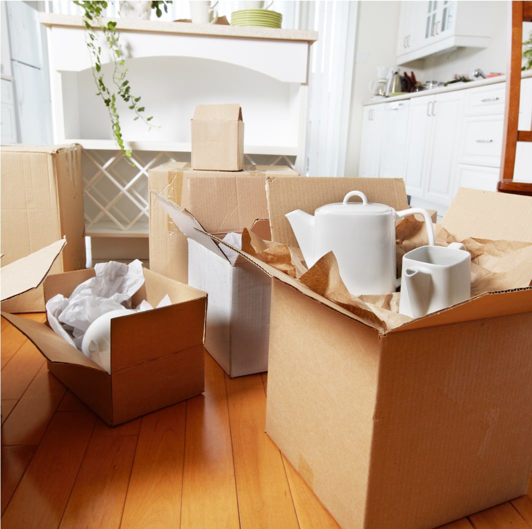 Several packed and unpacked cardboard boxes are scattered on a wooden floor in a brightly lit kitchen. Some boxes contain white kitchenware, including a teapot and cups. The kitchen in the background has white cabinets and a countertop with a few items on it.