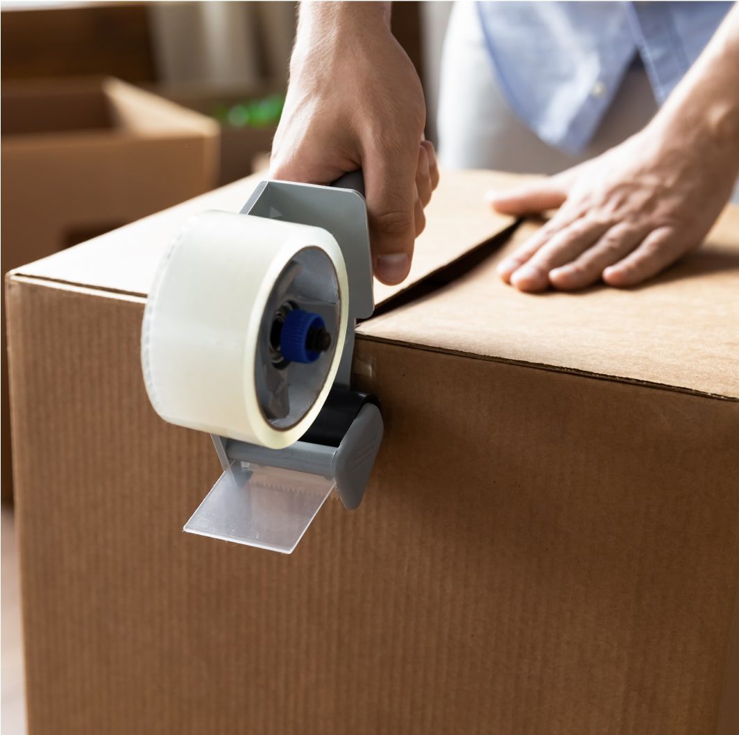 Close-up of a person sealing a cardboard box with clear packing tape using a tape dispenser. The box and hands are in focus, while other packing supplies are visible in the background.