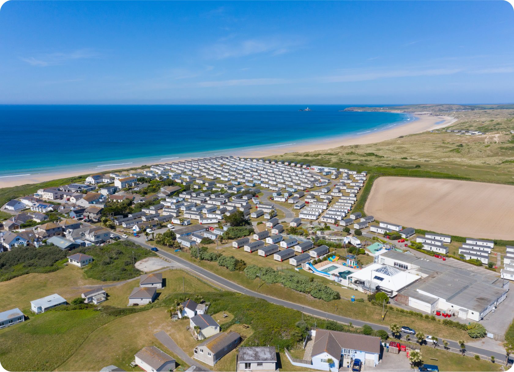 Aerial view of a coastal area with numerous mobile homes and buildings near a beach. The blue ocean and sandy shore stretch into the distance. The surrounding landscape includes fields, greenery, and a few scattered houses. The sky is clear and blue.