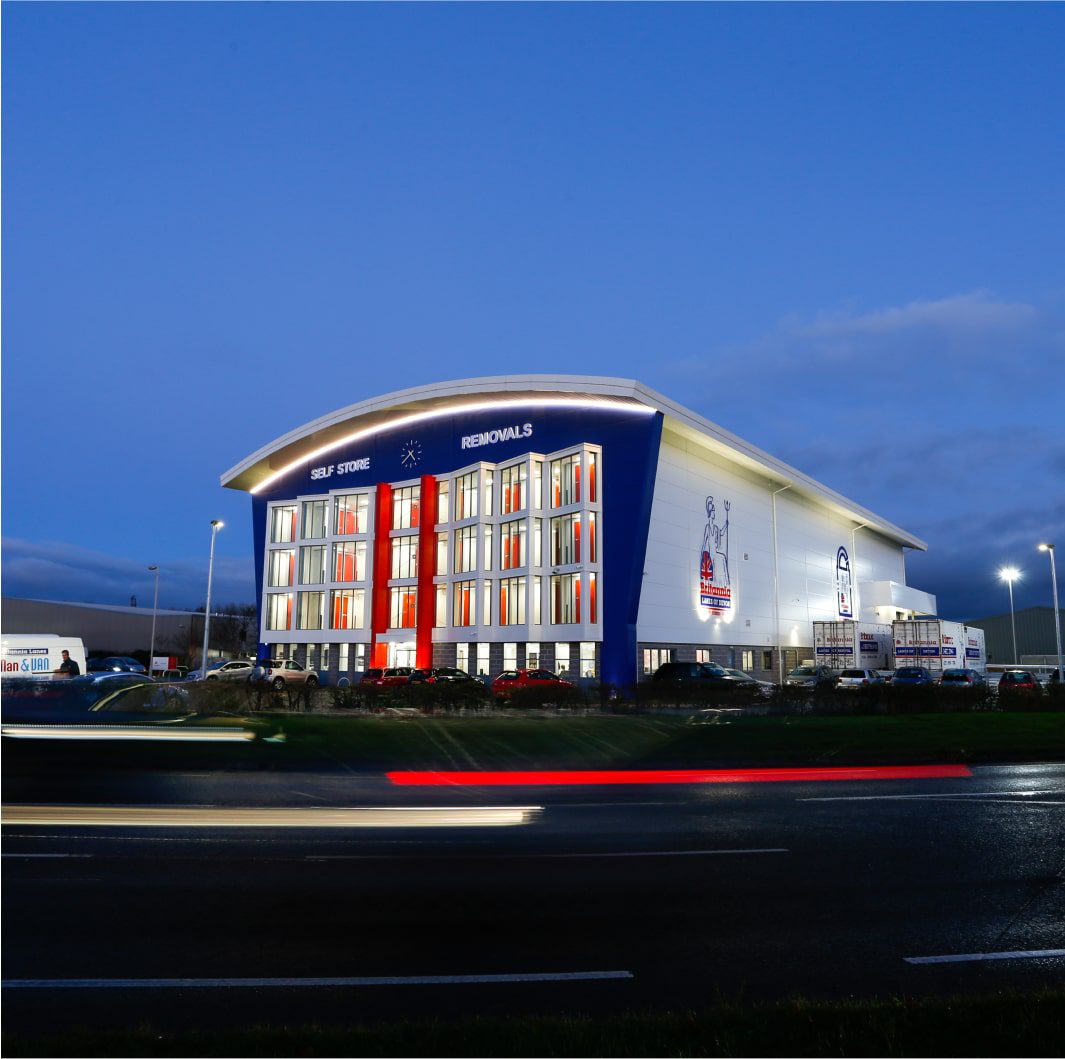 A modern, multi-story storage and removals facility is illuminated at dusk. The building has large glass windows and bright red accents. Several vehicles, including moving trucks, are in the foreground, with light trails from passing cars on the road in front.