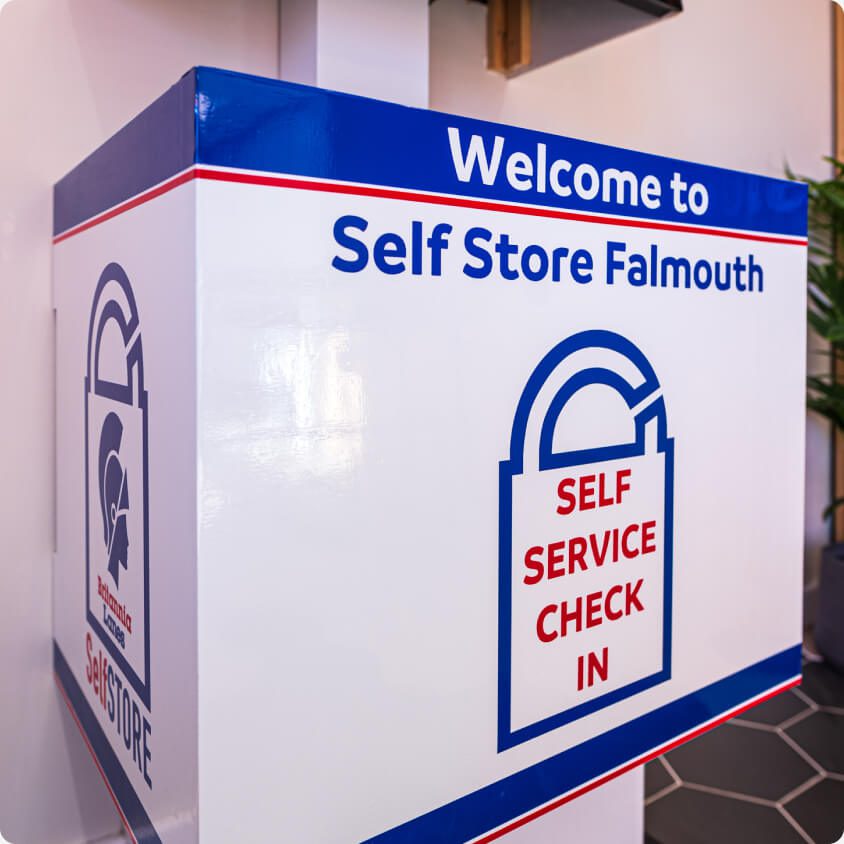 A white kiosk at Self Store Falmouth with blue and red trim. It features the text "Welcome to Self Store Falmouth" and "SELF SERVICE CHECK IN" with an image of a lock. The kiosk has a clean, modern appearance and is placed indoors.