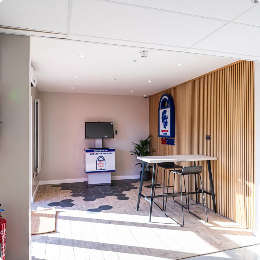 A modern, minimalist office space with light wood flooring and slatted wood paneling on one wall. It features a high table with stools, a wall-mounted screen, and a large branded sign. There is a potted plant and geometric patterned carpet tiles.