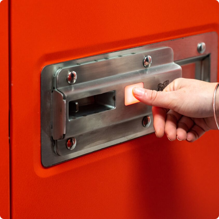 A hand pressing a white rectangular button on a metal panel mounted on an orange door. The panel appears to be part of a latch or locking mechanism.