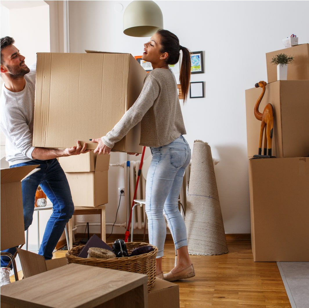 A man and a woman are moving a large cardboard box together in a room filled with packed boxes and furniture. The woman is smiling while lifting the box. There are various home items, including packed frames, a rolled-up rug, and a woven basket on the floor.