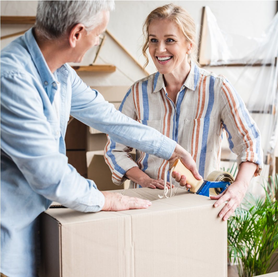 An elderly man and woman are packing a cardboard box. The man is holding the box while the woman is sealing it with packing tape, smiling at him. In the background, there are more boxes and some indoor plants.