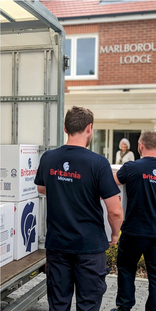 Two men wearing Britannia Movers T-shirts are unloading boxes from a moving truck outside a building labeled "Marlborough Lodge." One man is holding a box while the other stands beside him. An older woman stands in the background, watching the movers.