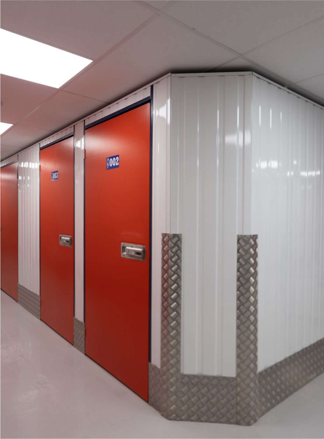 A brightly lit, clean storage facility corridor with orange doors numbered 002 and 003. The walls are lined with white panels and the lower section is protected by diamond plate metal. The floor is shiny and white. The ceiling has rectangular light panels.