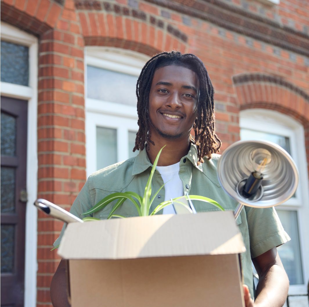 A person with dreadlocks stands in front of a brick house, smiling while holding a cardboard box containing a houseplant, a desk lamp, and other items, possibly indicating a move or new beginning.