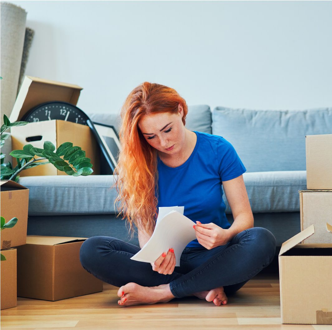 A person with long red hair sits on the floor in a casual outfit, looking at a piece of paper. They are surrounded by moving boxes and a potted plant, suggesting they are in the process of moving. A couch and a large clock can be seen in the background.