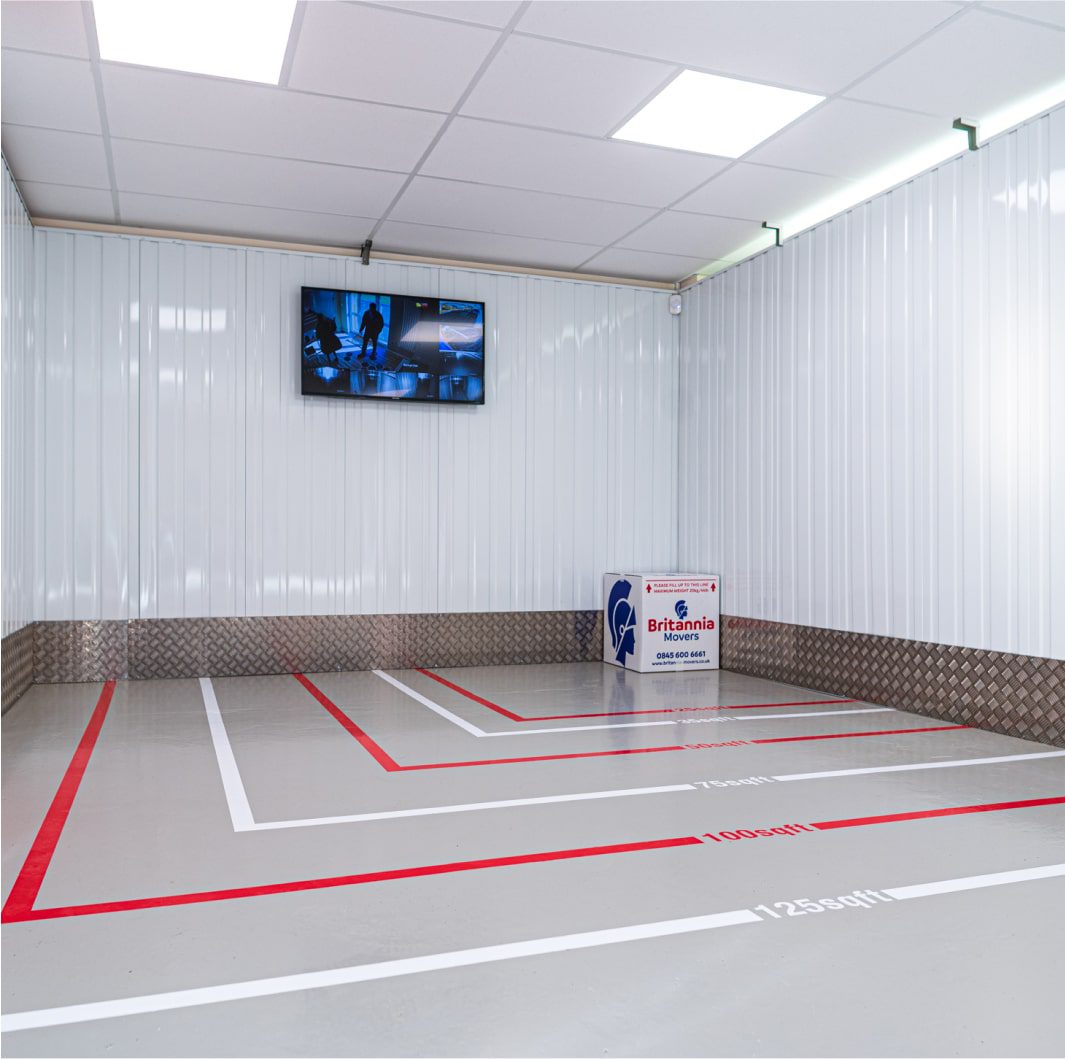 A clean, well-lit room with white corrugated walls and a grid-like pattern on the ceiling. The floor has red and white lines forming rectangular shapes with labels "1000N" and "12500N." A Britannia box sits on the floor, and a TV is mounted on the wall.