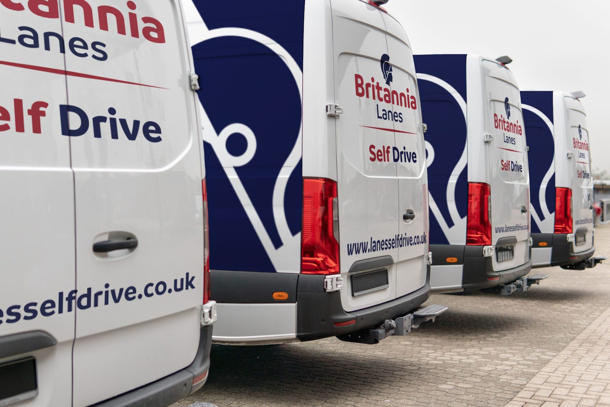 Several white vans are parked in a row, displaying the branding "Britannia Lanes Self Drive" with a logo on the rear, and the website "lanesselfdrive.co.uk" on the side. The background is a paved area, with a slightly cloudy sky overhead.