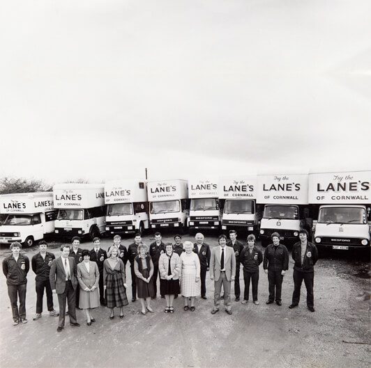 A black and white photo shows a group of 20 people lined up in front of seven trucks. The trucks display signs that read "Lane's of Cornwall." The group, comprised of men and women in formal and casual attire, stands posing for the camera on a clear day.