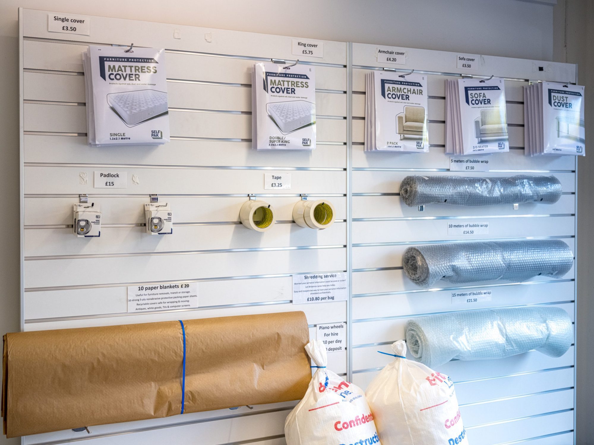 A store display showcasing various moving supplies, including mattress covers, armchair covers, and sofa covers hanging on a wall. Rolls of tape, packing bubble wrap, brown wrapping paper, and white padded bags are also visible. Prices are labeled for each item.