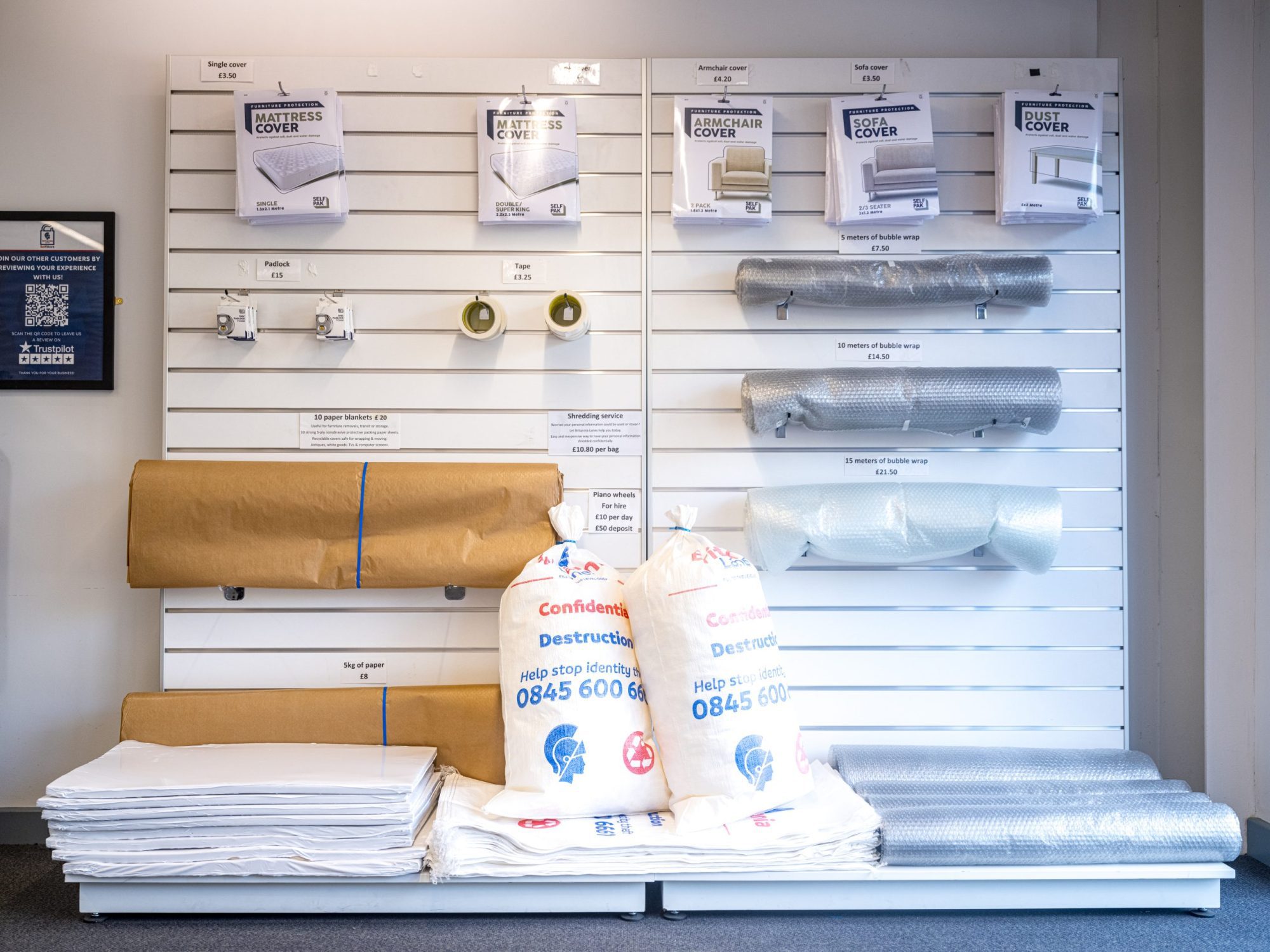 A neatly organized display shows various moving supplies, including mattress covers, sofa covers, bubble wrap, and packing paper. Two large bags filled with packing material and plastic wrap rolls are also placed on the floor in front of the display.