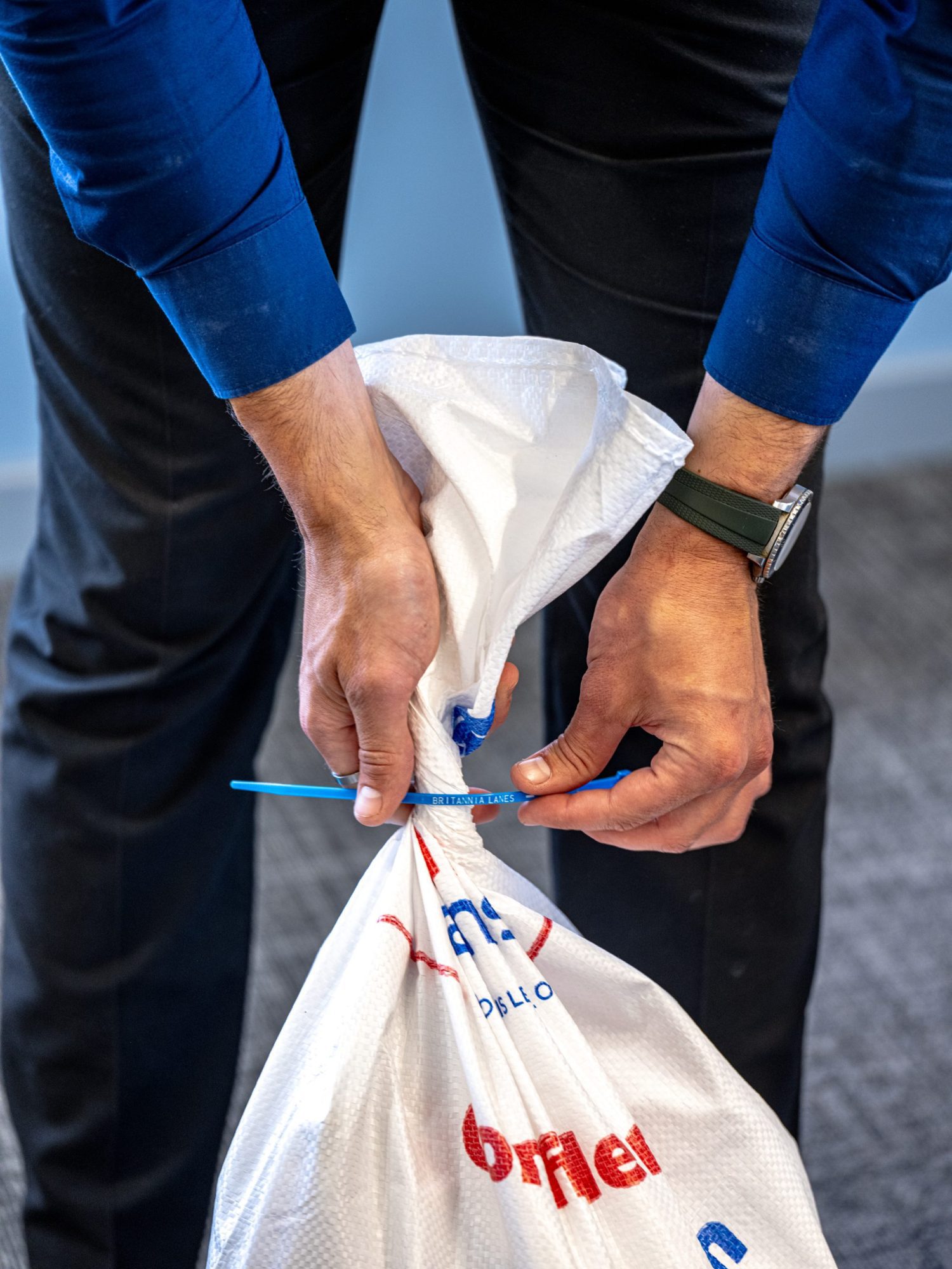 A person in a blue shirt and dark pants ties a white garbage bag with a blue zip tie. The bag features some red and blue text. The individual wears a green watch on their left wrist and stands on a carpeted floor.