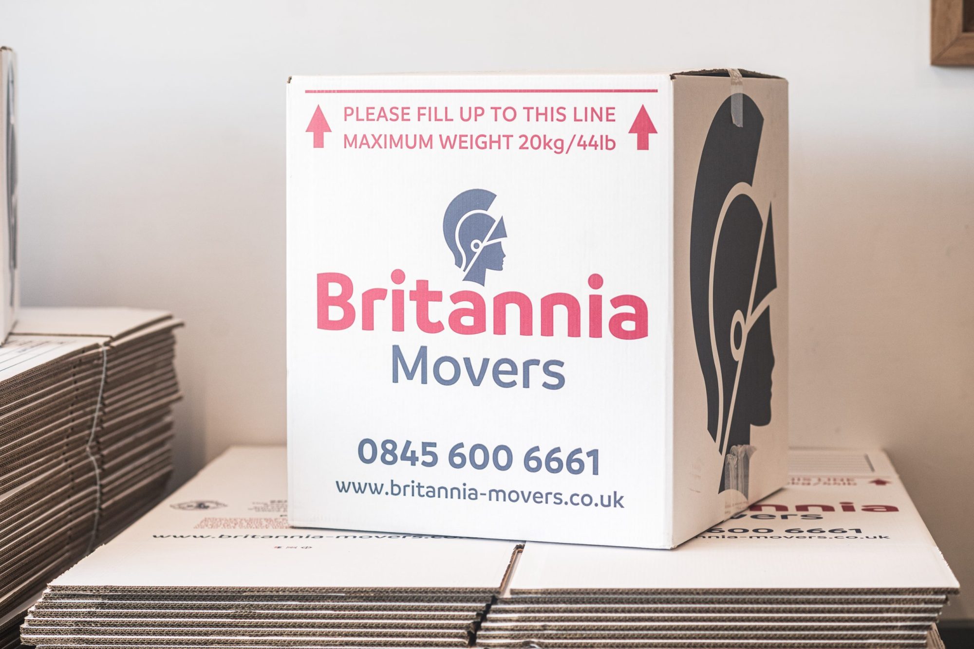 A stack of moving boxes from Britannia Movers, predominantly white with red and blue text. The box highlights a maximum weight limit of 20kg/44lb and the company's contact information, including a phone number (0845 600 6661) and website (www.britannia-movers.co.uk).