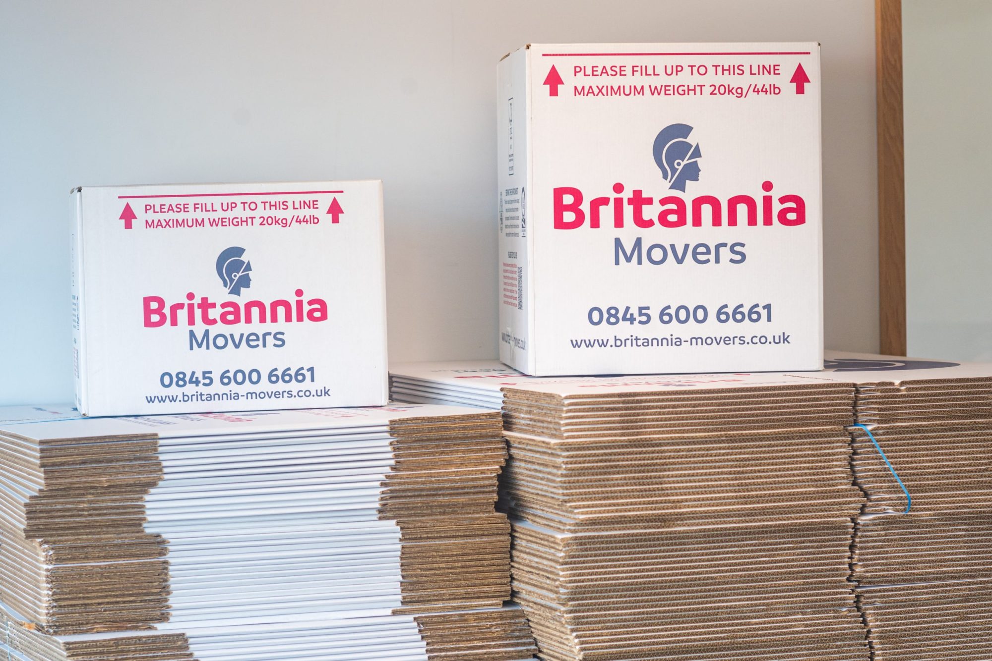 Two white boxes of different sizes with "Britannia Movers" branding are stacked on a pile of flattened cardboard boxes. The larger box has a red line indicating a maximum weight of 20 kg/44 lb. Contact details and website are displayed on both boxes.