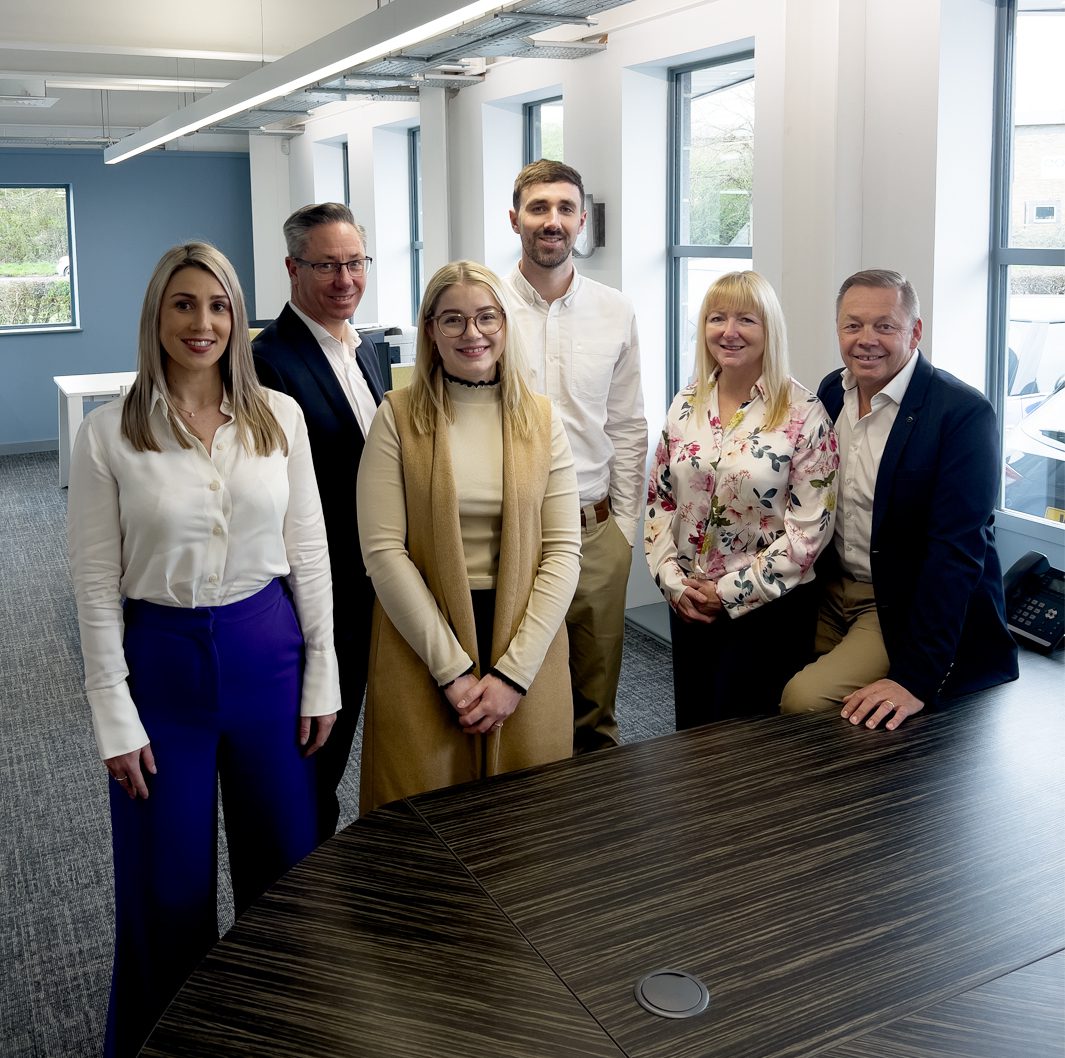 A group photo of five people in business attire standing and one person sitting on the edge of a conference table. They are in an office setting with large windows in the background. The group appears to be smiling and posing for the camera.