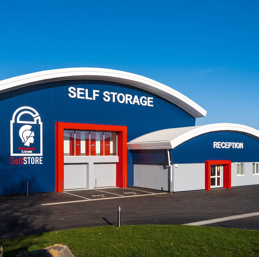 A blue and white self-storage facility with large signage reading "SELF STORAGE" and "RECEPTION." The building has red accents around the entrances and a logo featuring a lock and helmeted figure. The area is paved with a clear, blue sky above.