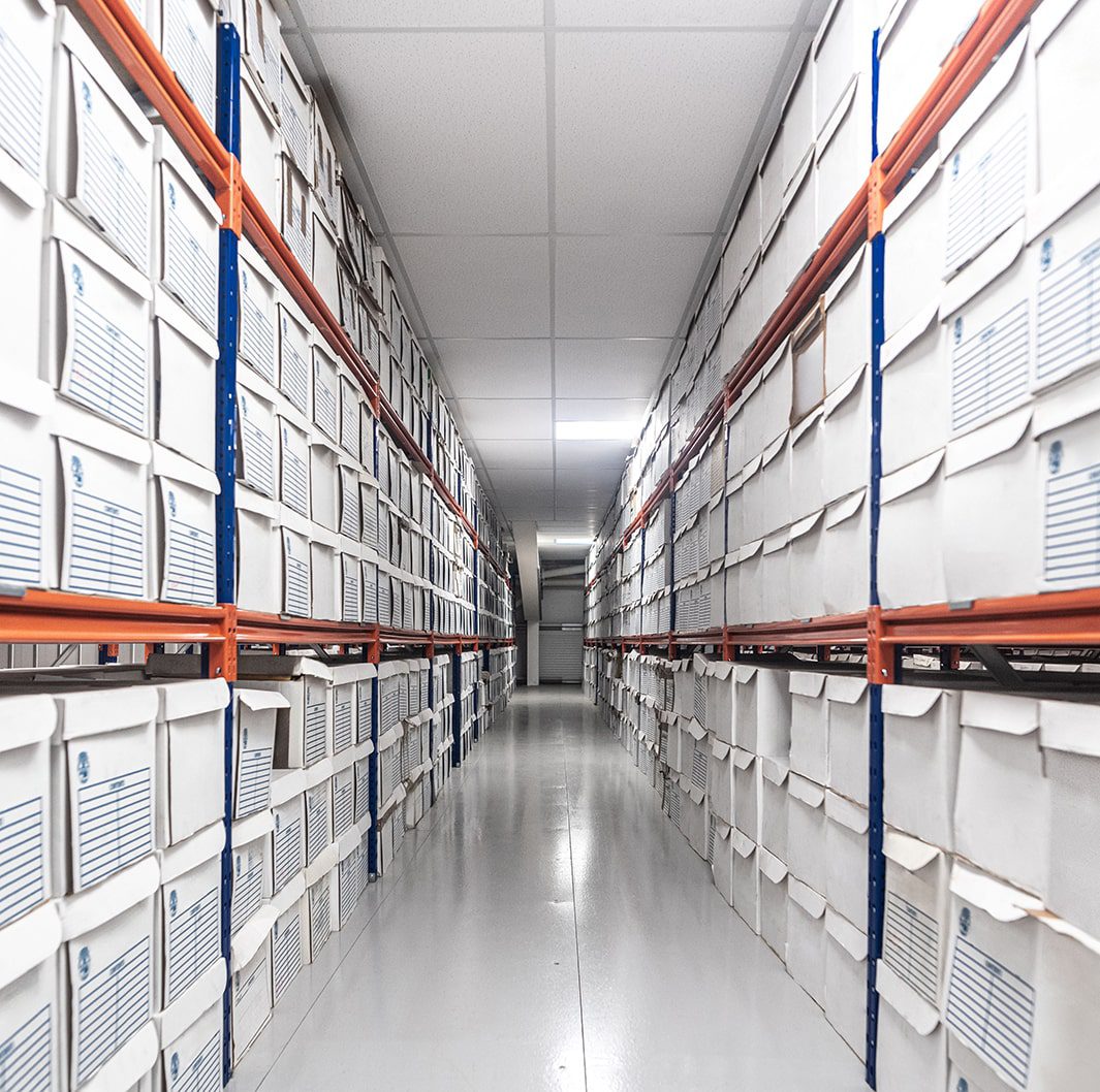 A long hallway filled with rows of white storage boxes on metal shelves, extending into the distance. The boxes have blue labels and are neatly arranged, creating a tidy and organized environment. The floors are polished and the ceiling has white tiles.