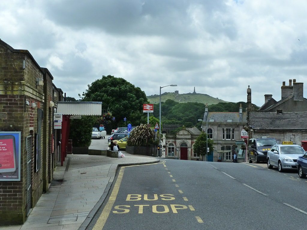 A street view in a small town with a "Bus Stop" sign painted on the road. Buildings line the street, and cars are parked beside the road. Trees and a hill with a monument are visible in the background under a cloudy sky.